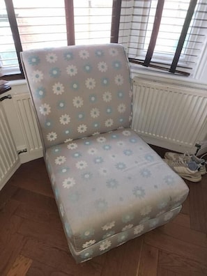 Una butaca en esquina con estampado floral, ubicada junto a un radiador y un par de zapatos.