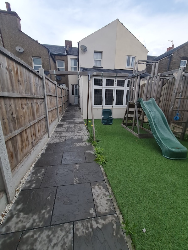 Un patio trasero con un sendero pavimentado, césped artificial, una cerca de madera y un tobogán verde.