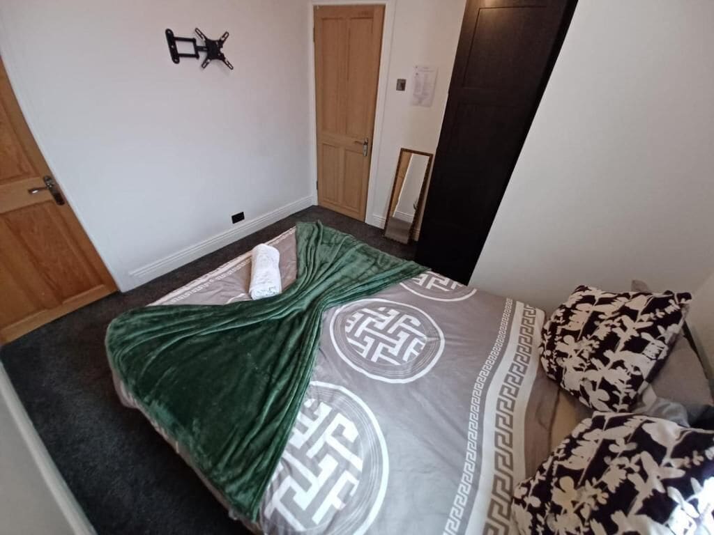 Un dormitorio con cama, manta verde, almohadas estampadas, puerta de madera y un espejo en la pared.