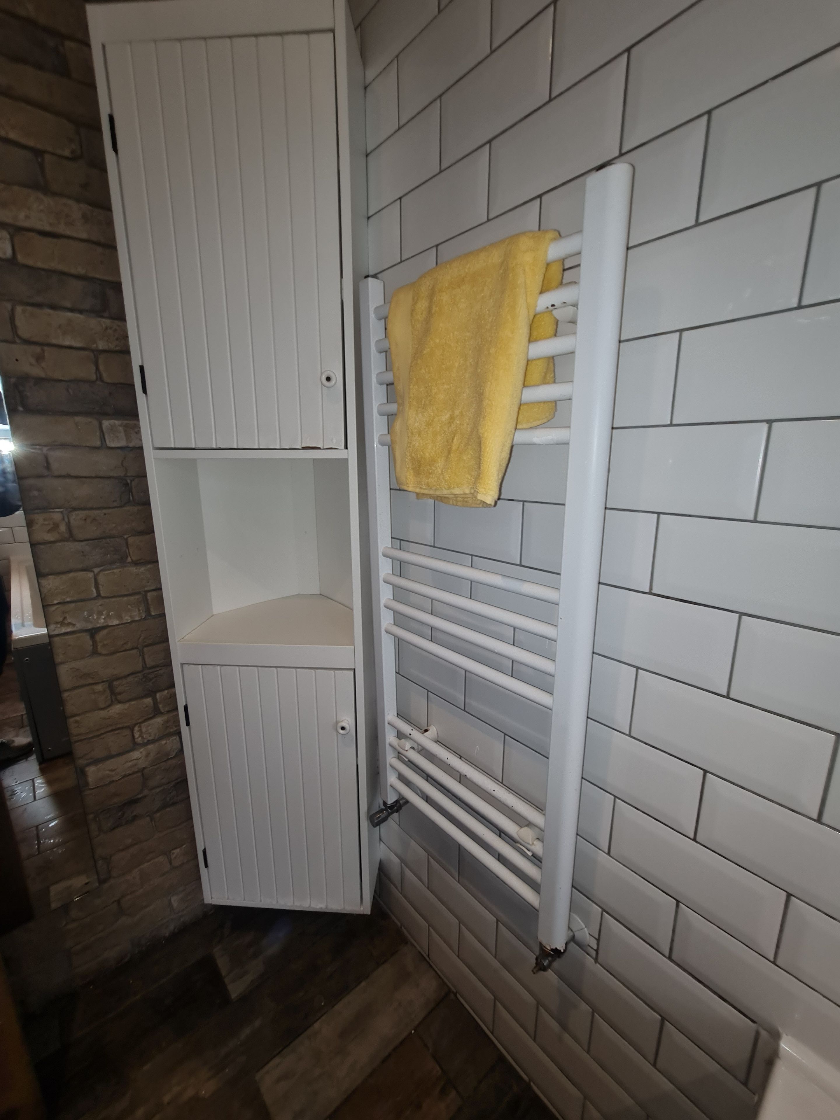 Un baño con pared de azulejos blancos, suelo de madera, un toallero blanco y una toalla amarilla, además de un armario blanco.