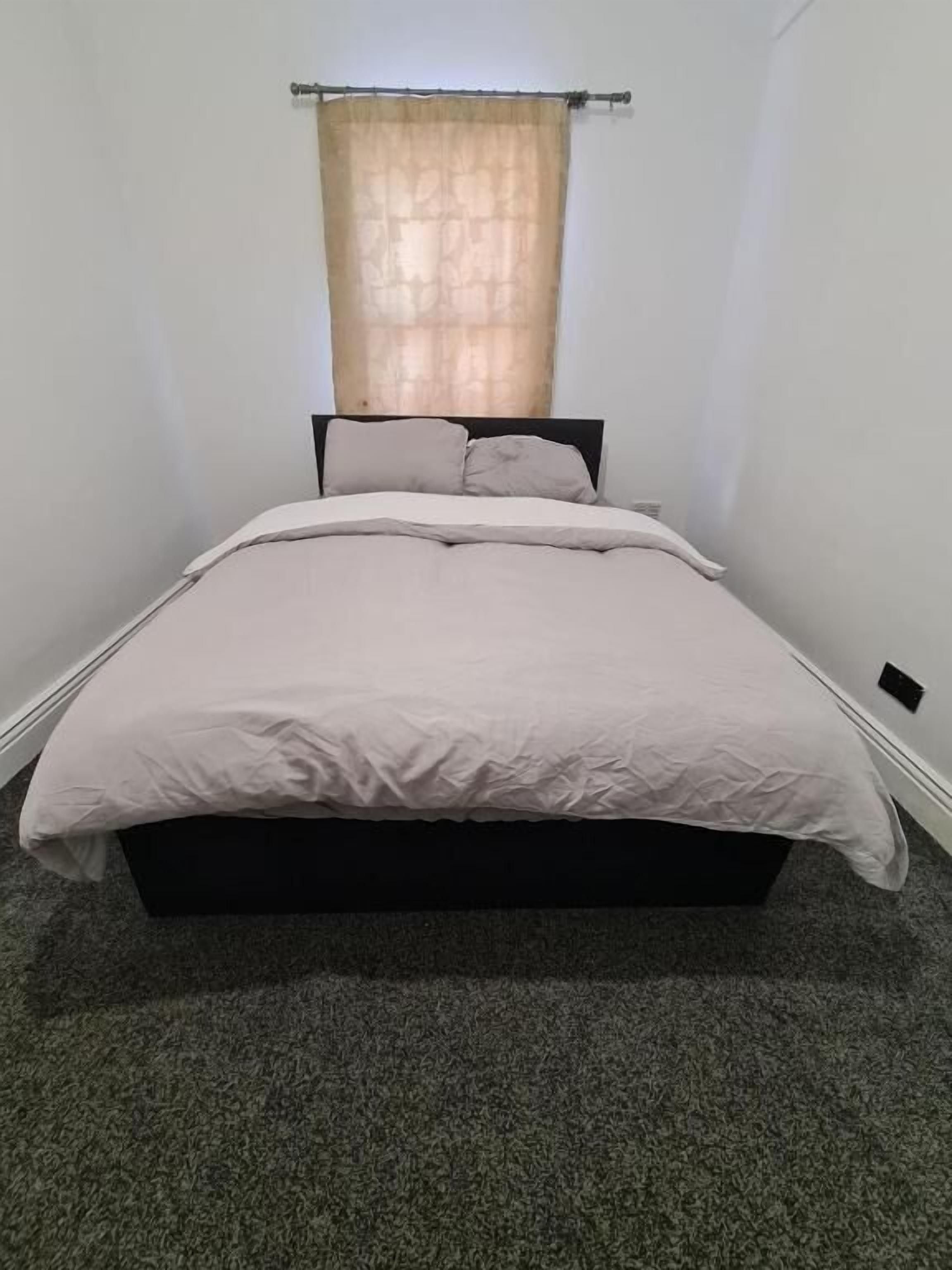 A single bed with a dark frame, a light-colored bedspread, and a window with a sheer curtain.
