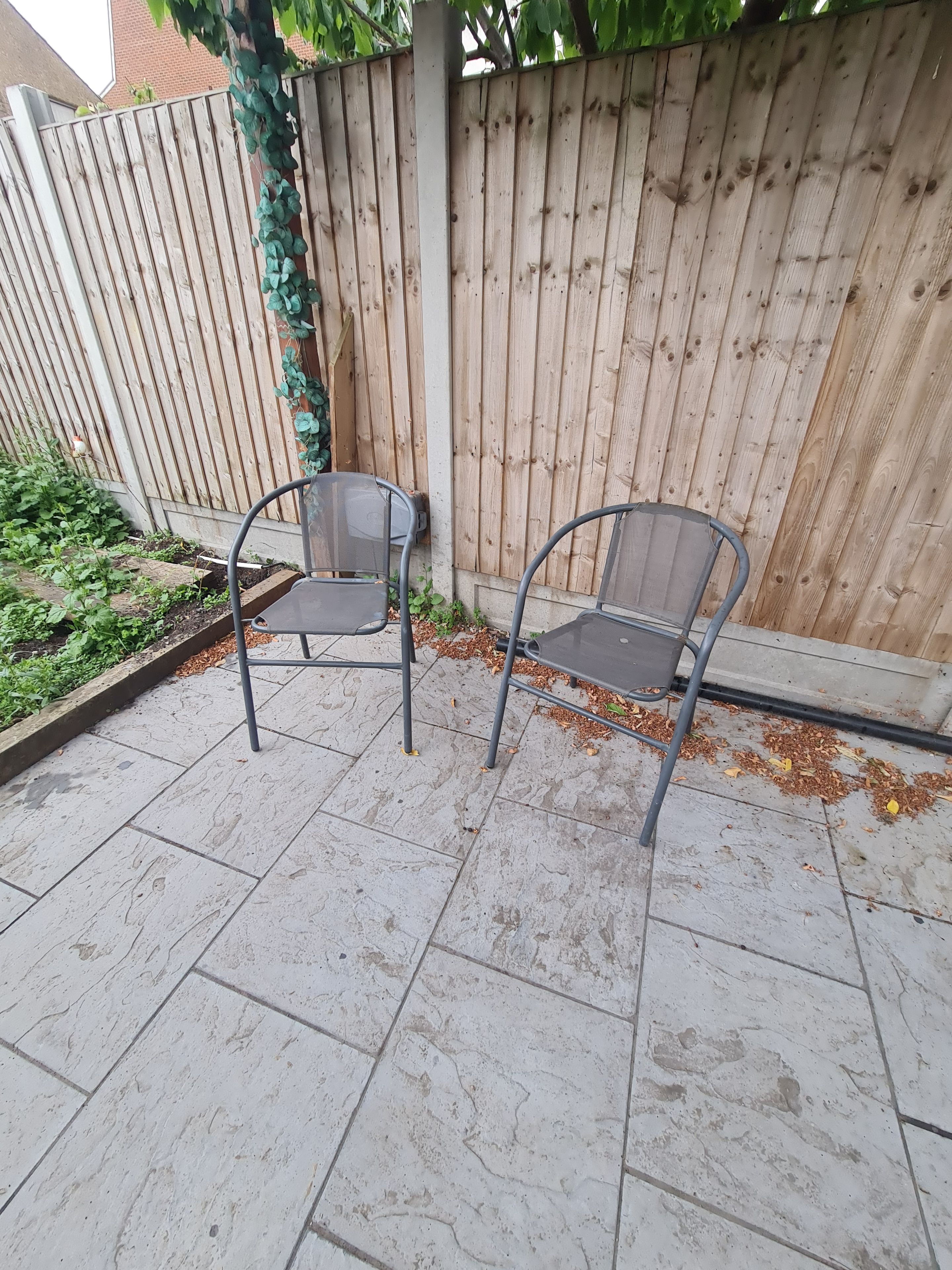 Dos sillas de metal en un patio con baldosas, una valla de madera y follaje al fondo.