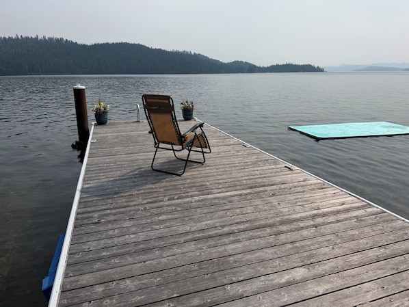 Un muelle de madera que se adentra en aguas tranquilas, con una silla plegable y plantas en macetas.