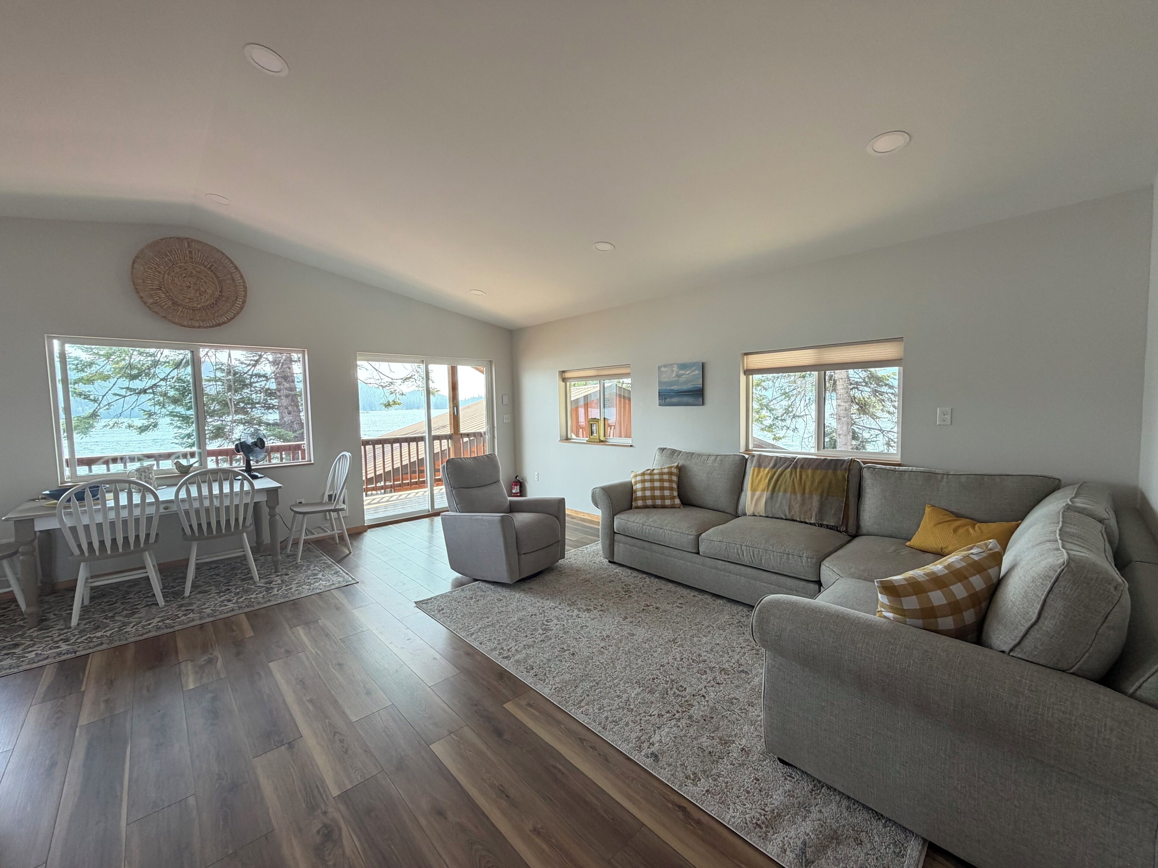 Ein geräumiges Wohnzimmer mit grauem Sofa, einem Esstisch mit Stühlen und großen Fenstern mit Blick ins Freie.