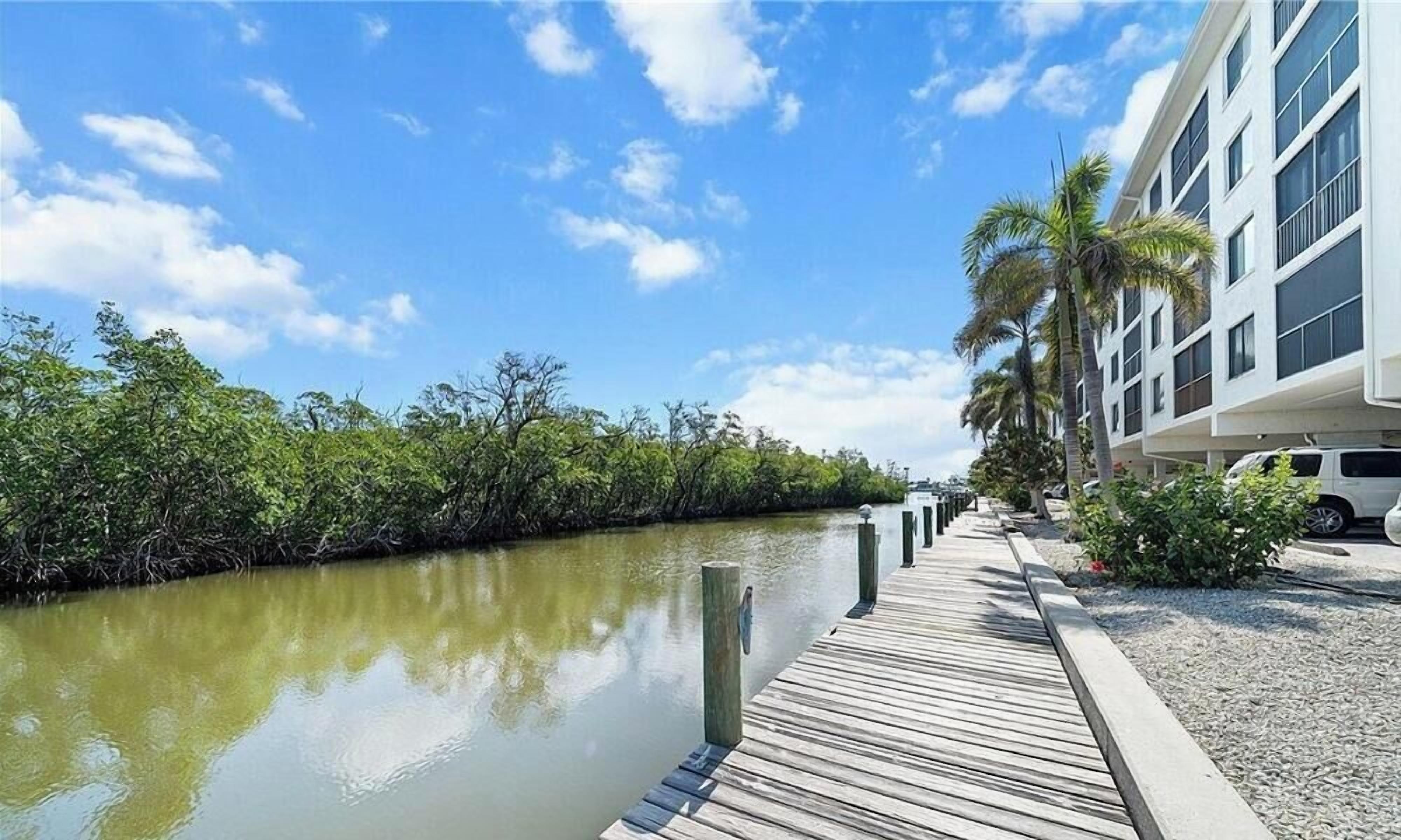 A waterfront hotel with a wooden dock, palm trees, and a clear sky.