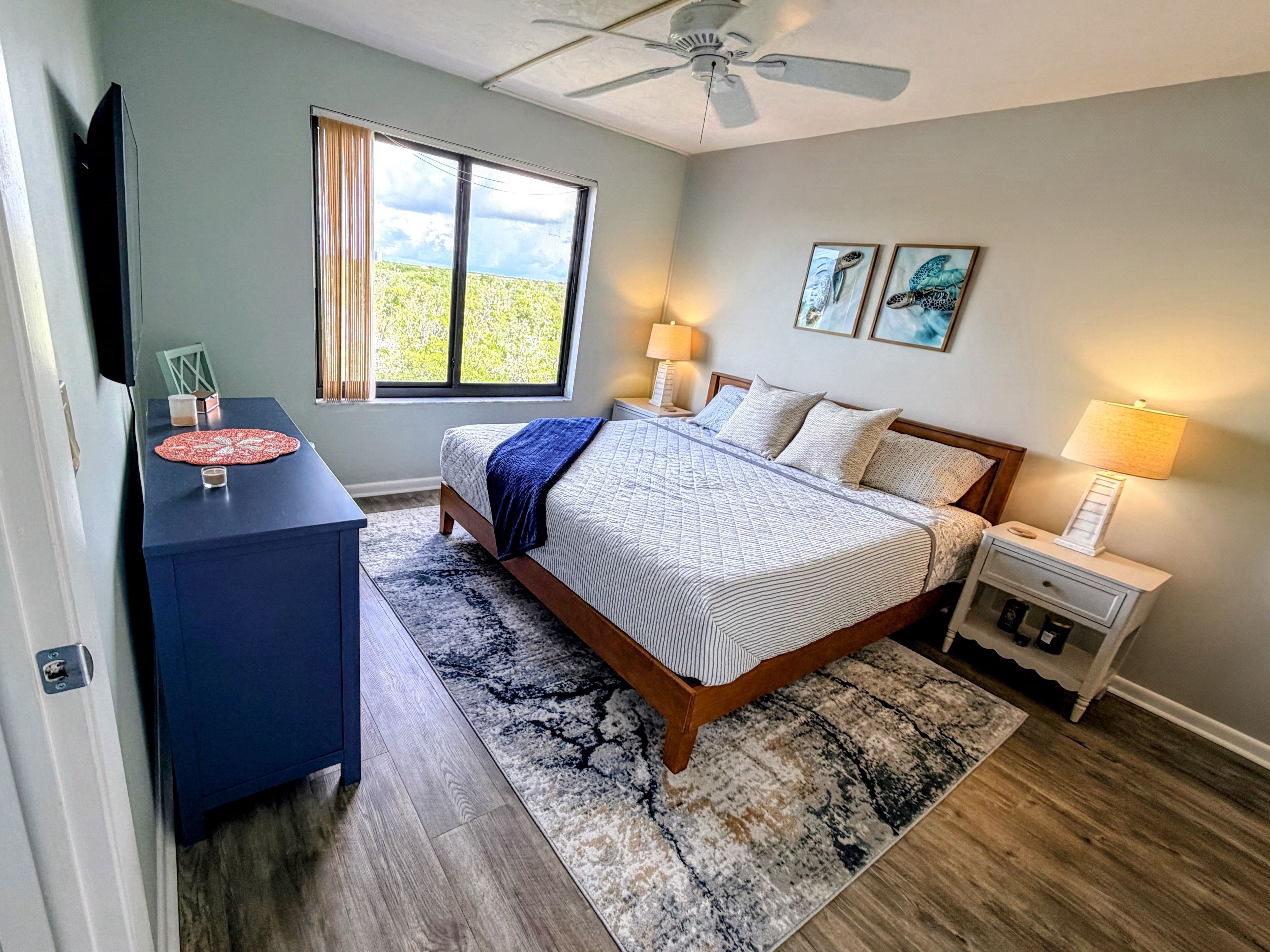 A bedroom with a bed, bedside tables, a desk, a ceiling fan, a window with curtains, and artwork on the wall.