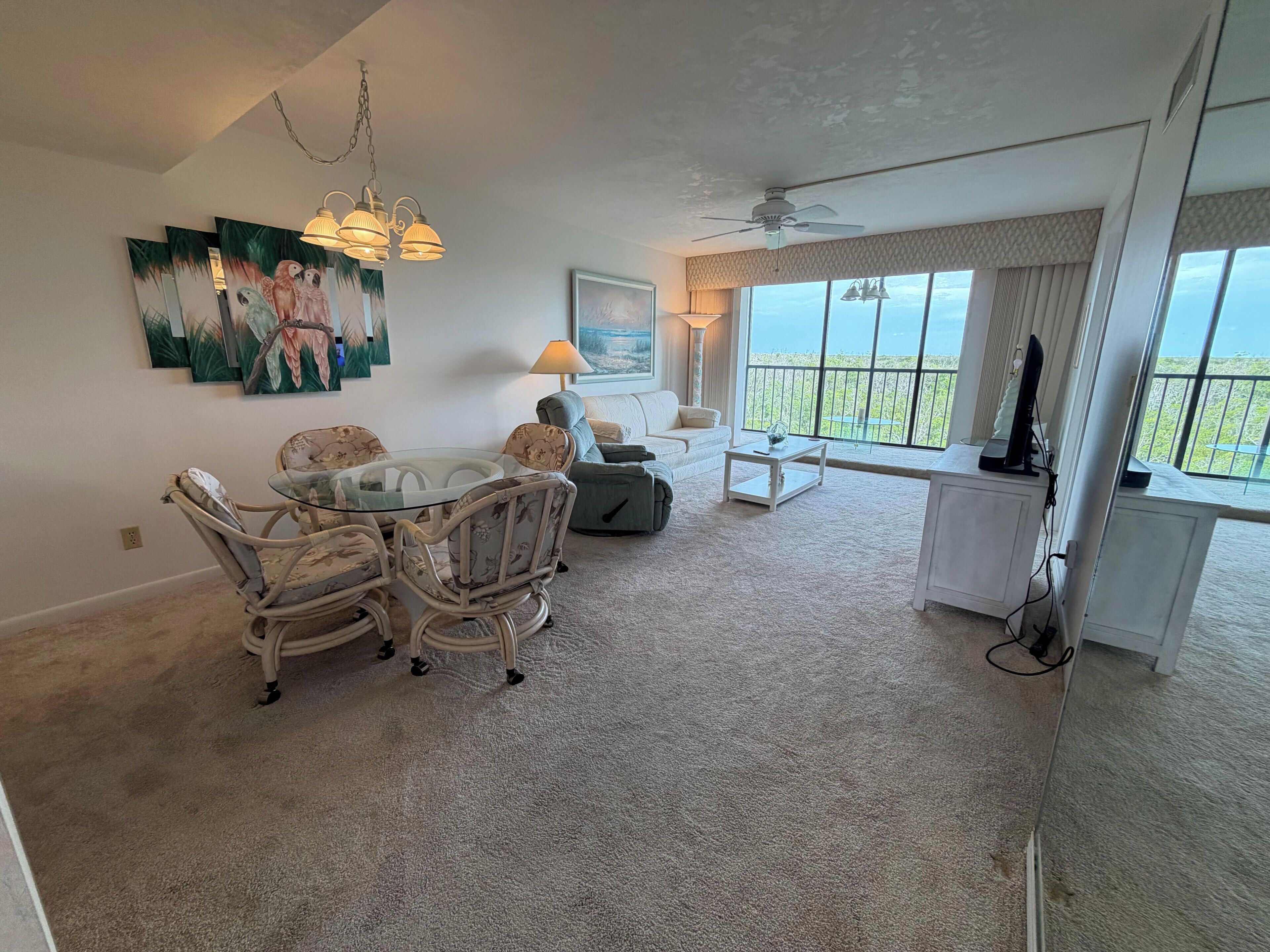 A living room with a glass dining table, white sofas, a ceiling fan, and a large window with a view.