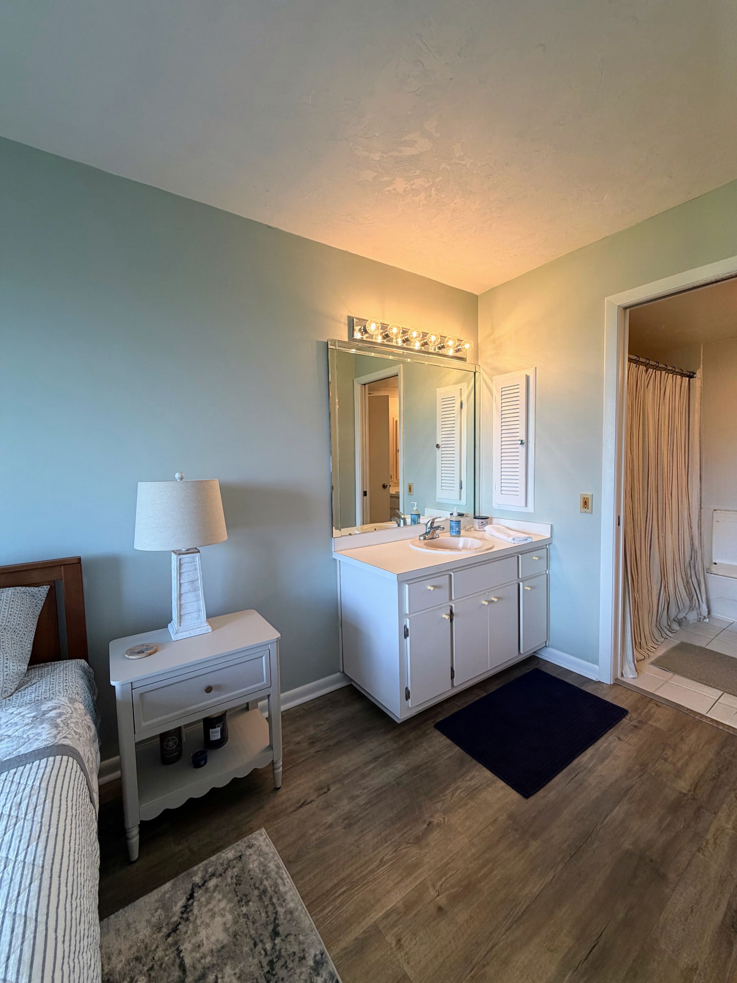 Un dormitorio con cama, mesita de noche con lámpara, tocador con espejo e iluminación, y una puerta que da a un baño.