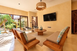 Een ruime zithoek met een ronde houten tafel, twee comfortabele fauteuils, een flatscreen-tv aan de muur en een groot raam met uitzicht op een zwembad en groen.