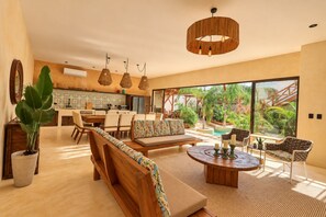 A spacious living area with a large window overlooking a pool and garden, wooden furniture, and a modern kitchen in the background.