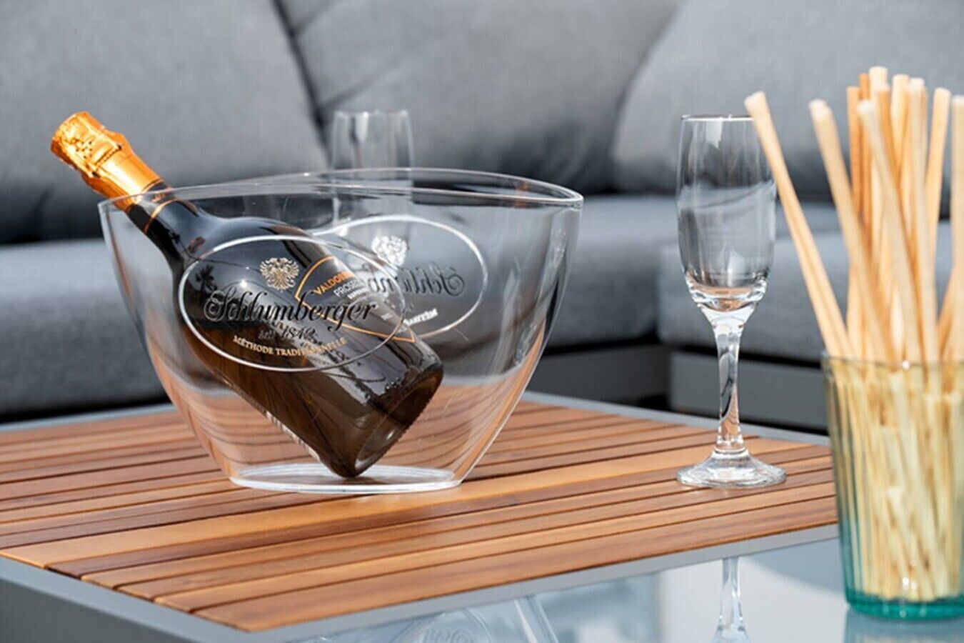 A clear glass champagne bottle with a label, resting on a glass bowl with a logo, next to a glass and a container of wooden sticks on a table.