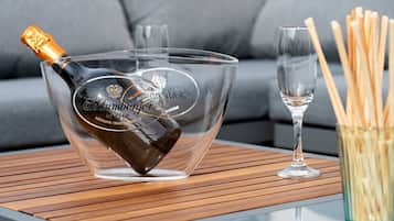 Eine klare Champagnerflasche mit Etikett, auf einer Glasbowl mit einem Logo platziert, neben einem Glas und einer Holzzündhölzchen-Box auf einer Tischplatte.