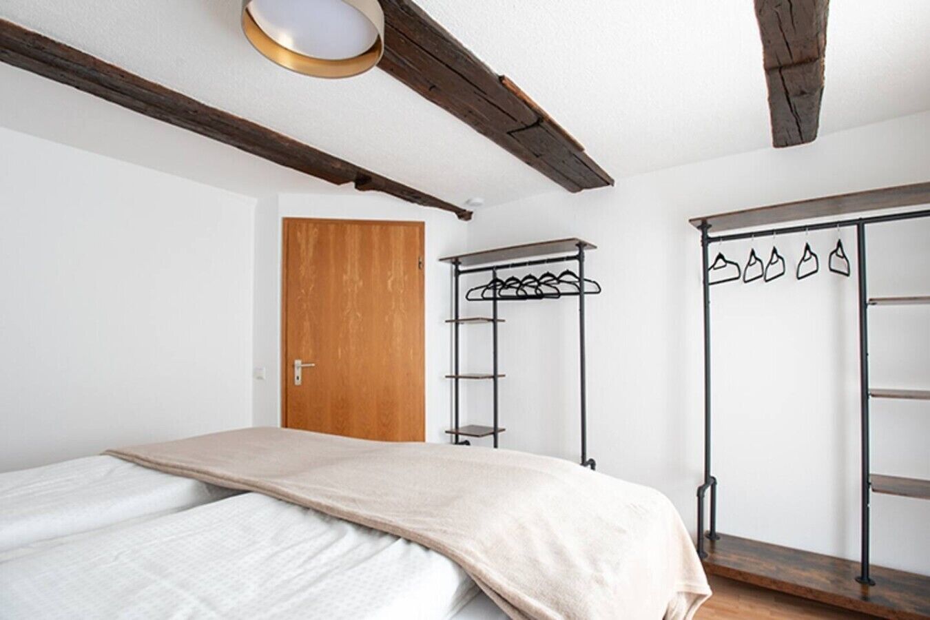 A bedroom with a bed, a wooden door, a metal coat rack, and a wardrobe with hanging items.