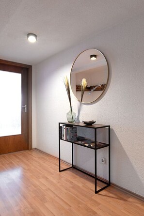 A room with a wooden floor, a console table with books and a vase, a round mirror on the wall, and a door with a wooden frame.