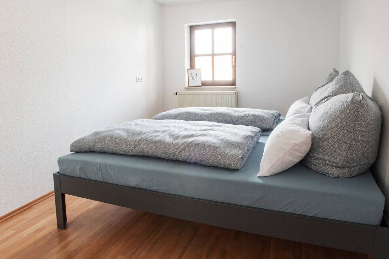 Ein Einzelbett mit grauer Matratze und Kissen, ein Fenster mit Jalousien und ein Heizkörper darunter.