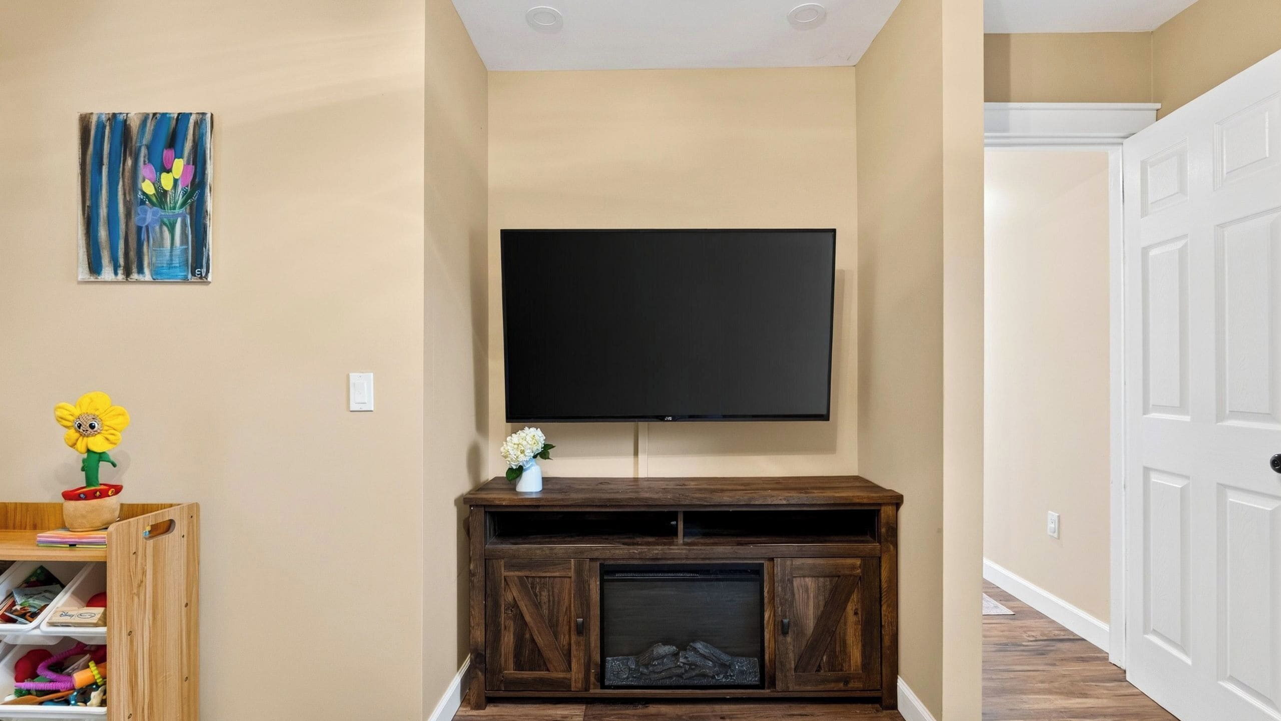 Uma sala de estar com uma TV de tela plana presa a um móvel de madeira, um pequeno vaso branco com flores e um quadro colorido na parede.