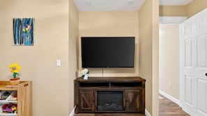 Uma sala de estar com uma TV de tela plana presa a um móvel de madeira, um pequeno vaso branco com flores e um quadro colorido na parede.