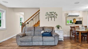 A living room with a grey sectional sofa, a dining area with wooden chairs, a staircase, and a wall-mounted tree decoration. - Ohana Nature Escape by Avantstay Creekside + Firepit + Grill Waterfall + Beaver Pond Walks (Catskill)