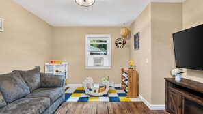 A living room with a grey sofa, a white play area, a wooden bookshelf, a television, and a window with a view of greenery. - Ohana Nature Escape by Avantstay Creekside + Firepit + Grill Waterfall + Beaver Pond Walks (Catskill)