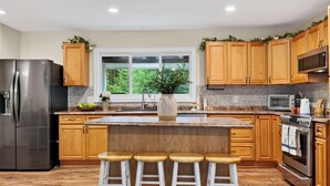 A modern kitchen with wooden cabinets, a stainless steel refrigerator, a large island with stools, and a view of greenery outside. - Ohana Nature Escape by Avantstay Creekside + Firepit + Grill Waterfall + Beaver Pond Walks (Catskill)