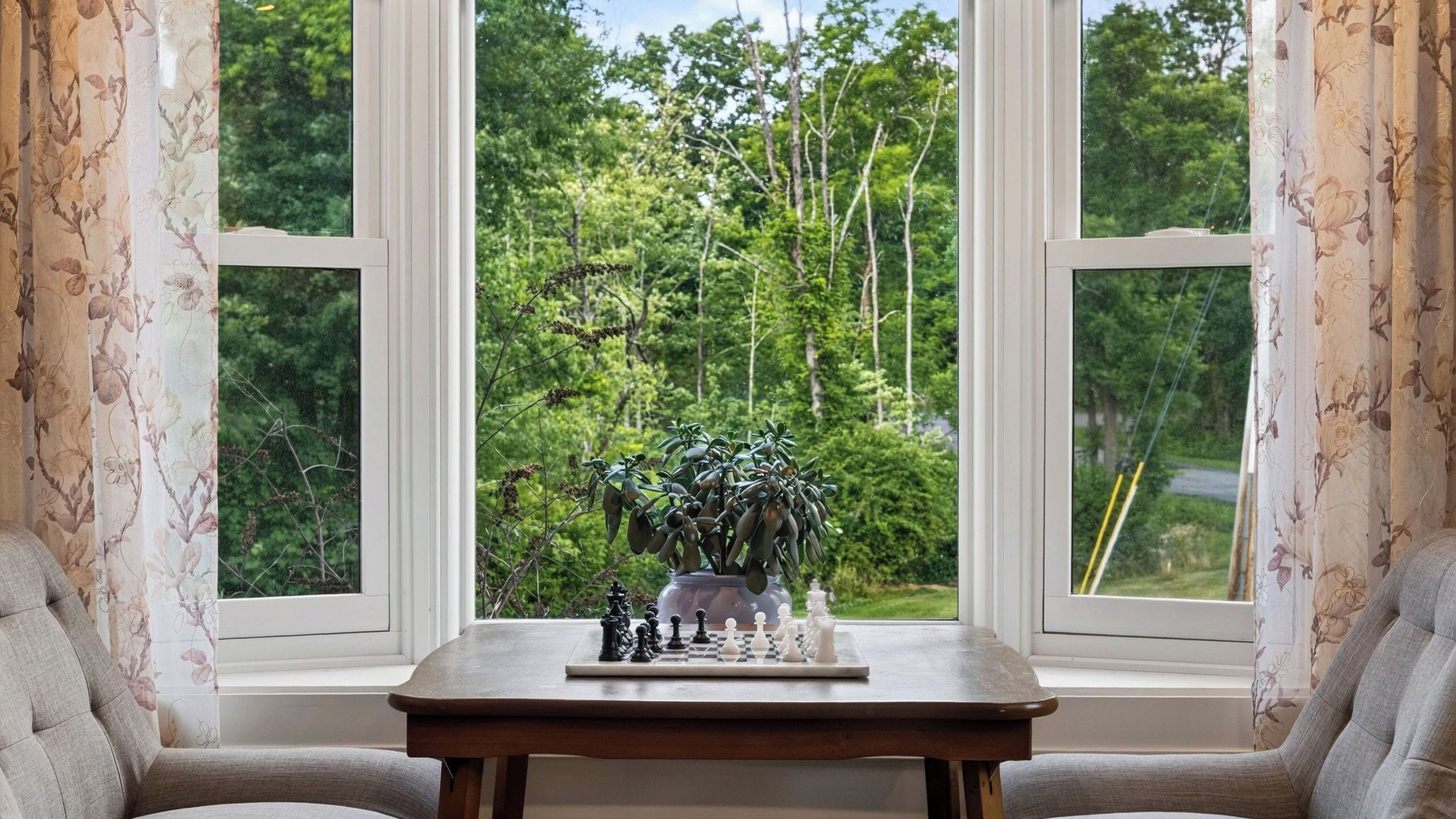 A chessboard on a table by a window with floral curtains, overlooking a forest.