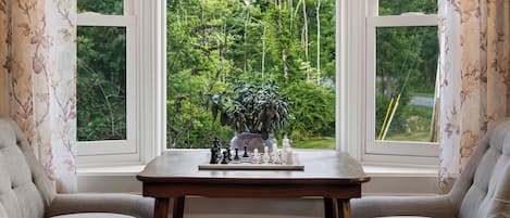 A chessboard on a table by a window with floral curtains, overlooking a forest.