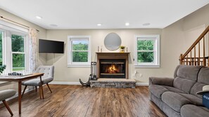 A living room with a fireplace, a television mounted on the wall, and a staircase leading to another level. - Ohana Nature Escape by Avantstay Creekside + Firepit + Grill Waterfall + Beaver Pond Walks (Catskill)