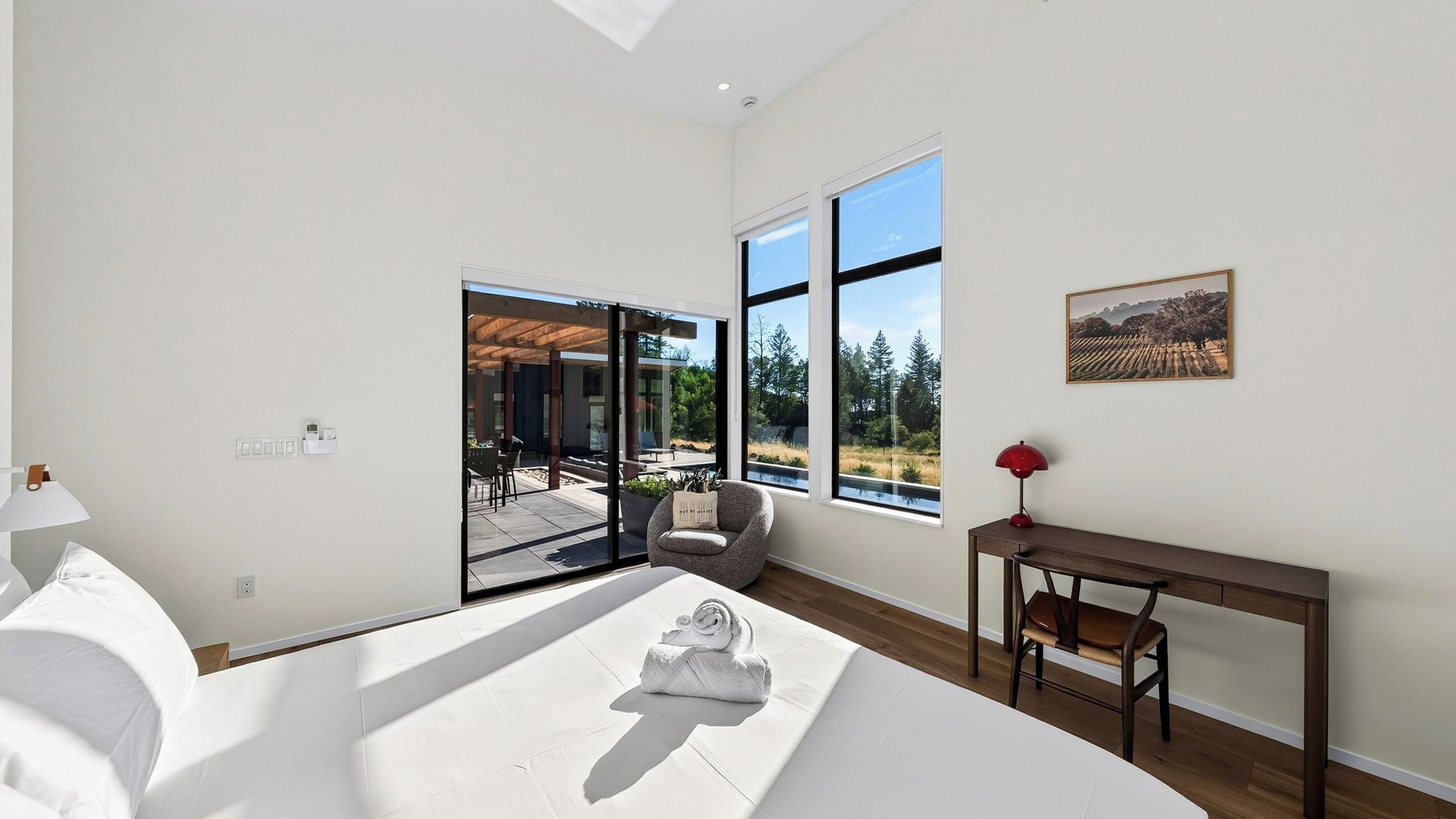 A modern bedroom with a large bed, a seating area with a chair and a table, and a view of a garden through large windows.