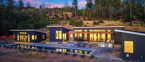 Una casa moderna con piscina, mobiliario exterior y vistas a un promontorio boscoso.