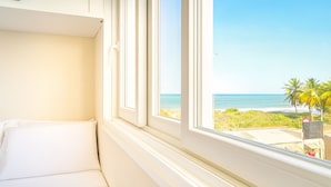 Una habitación con una cama blanca, una ventana con vista a la playa y palmeras.