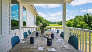 A wooden table set for dining with chairs, a vase with flowers, and cans of beverage on a porch with a view of a green landscape.
