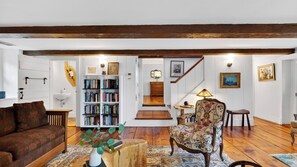 A spacious living room with a wooden floor, a sofa, a chair, a bookshelf, and a staircase leading to another level. - Ashokan Acres by Avantstay Private 125-acre Retreat Pond + Hot Tub + Sauna + Trails (Kingston)