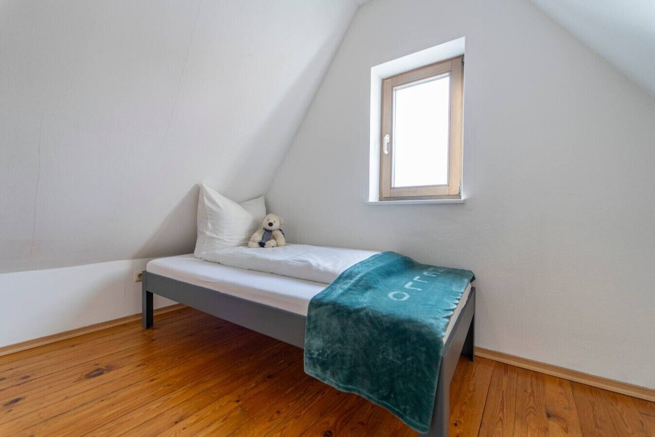 Una habitación pequeña con una cama individual, techo inclinado, ventana y piso de madera.