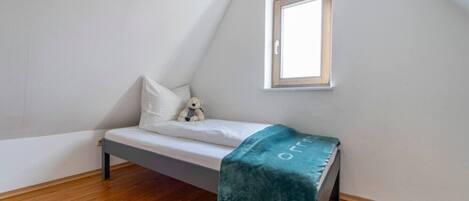 A small, single-bed room with a sloped ceiling, a window, and a wooden floor.