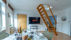 Un coin repas avec une table dressée pour deux personnes, un téléviseur fixé au mur et un escalier en bois.