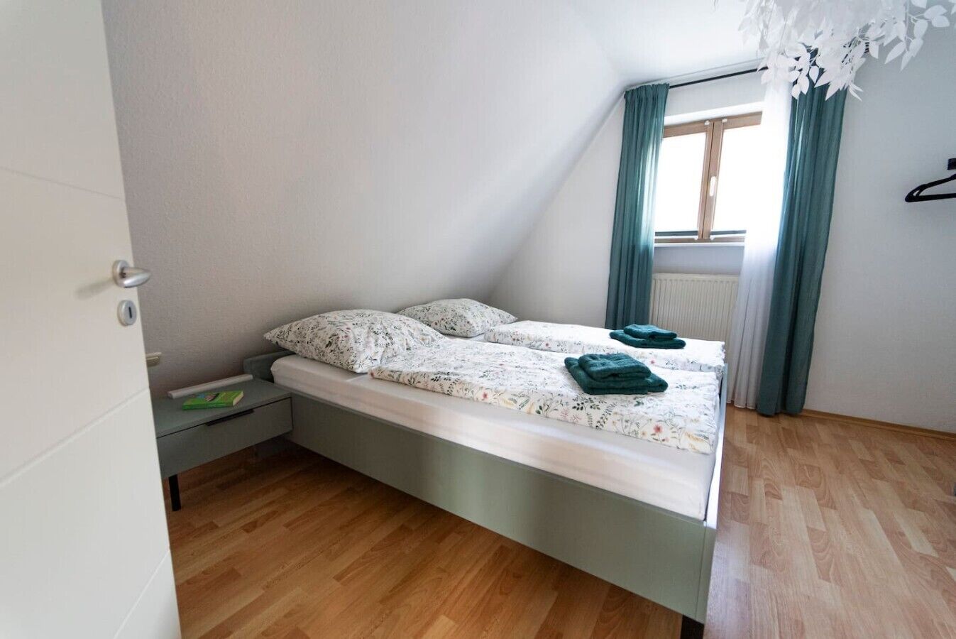 Una cama individual con una colcha estampada y dos almohadas, suelo de madera, una ventana con cortinas y un radiador.