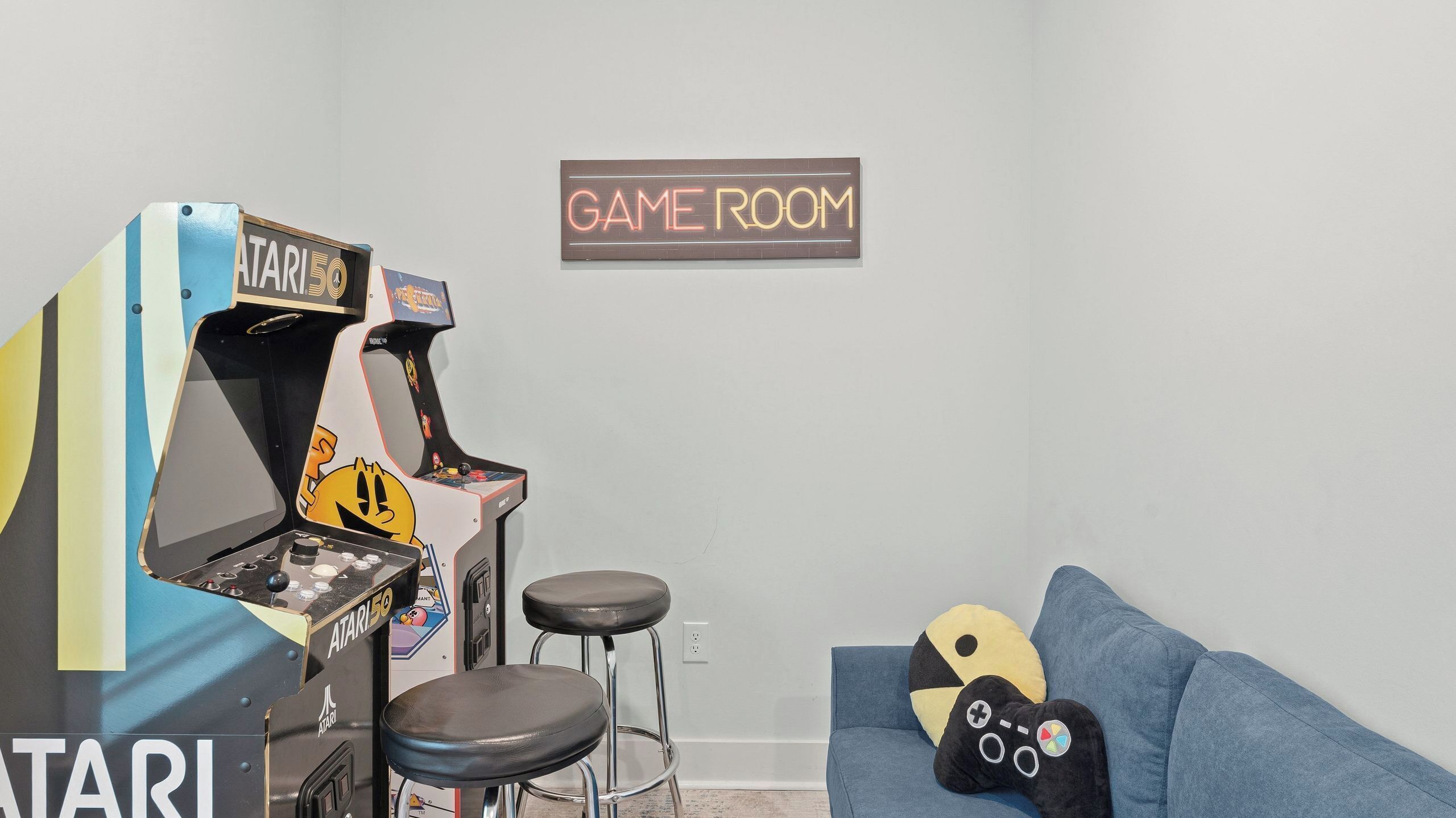 Une pièce équipée d’une console de jeux, de deux tabourets, d’un canapé et d’une enseigne murale indiquant « SALLE DE JEUX ».