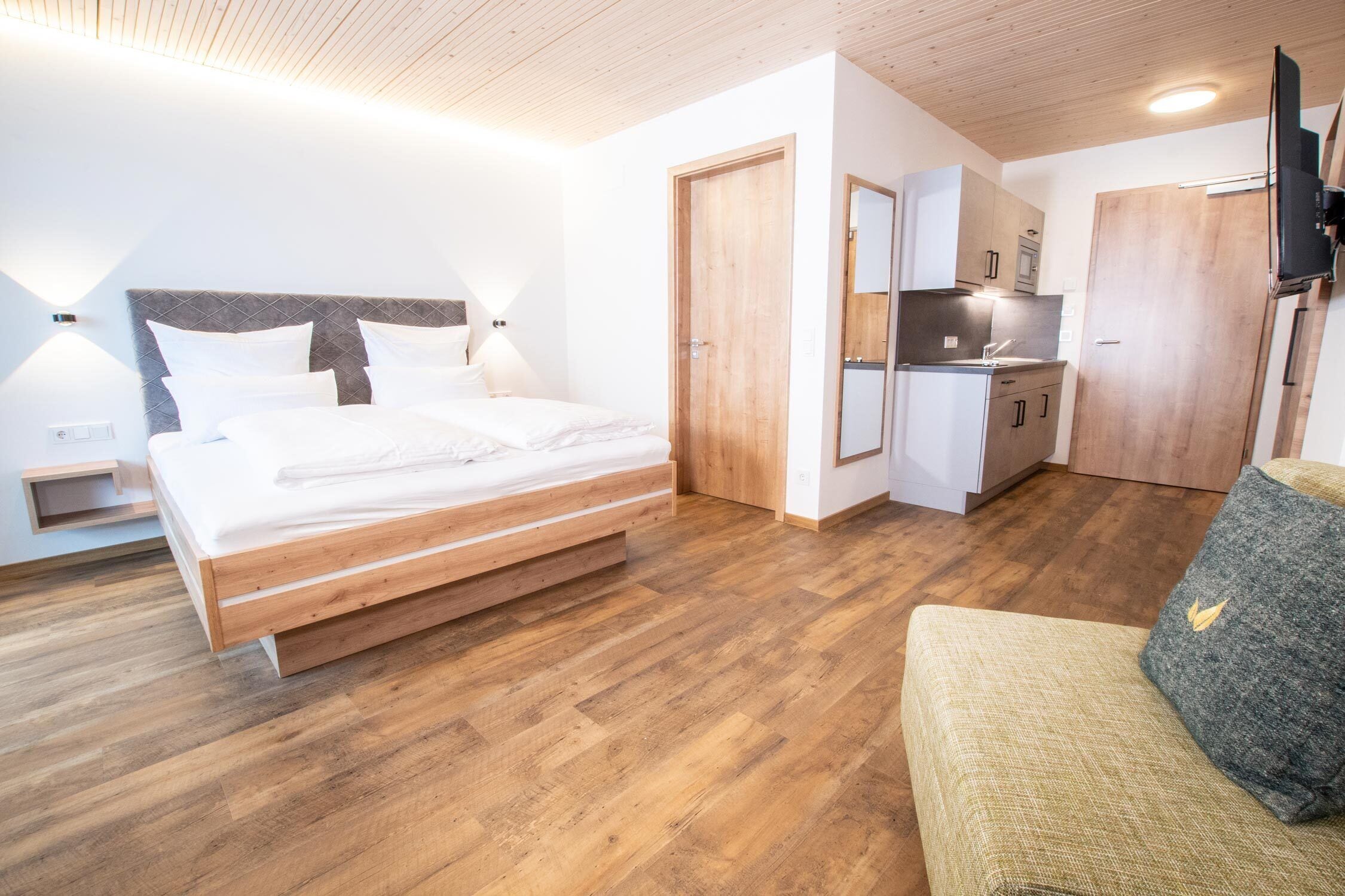 A modern hotel room with a wooden bed, a kitchenette, and a seating area.