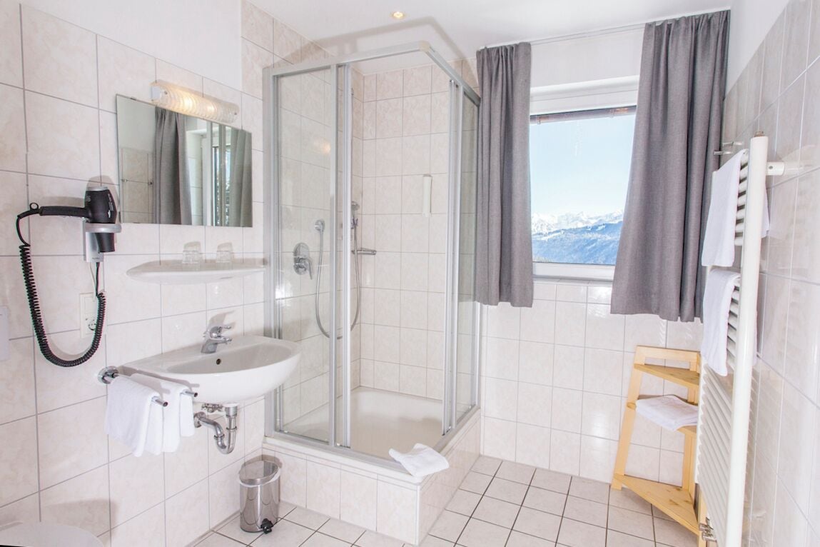 Ein Badezimmer mit einer Duschkabine, einem Waschbecken, einem Fön und einem fensterförmigen Ausblick mit Vorhängen.