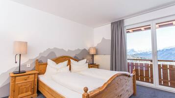 Ein Schlafzimmer mit einem hölzernen Bett, zwei Nachttischen, Blick auf Berge durch ein großes Fenster und ein Balkon mit Holzbalkon.