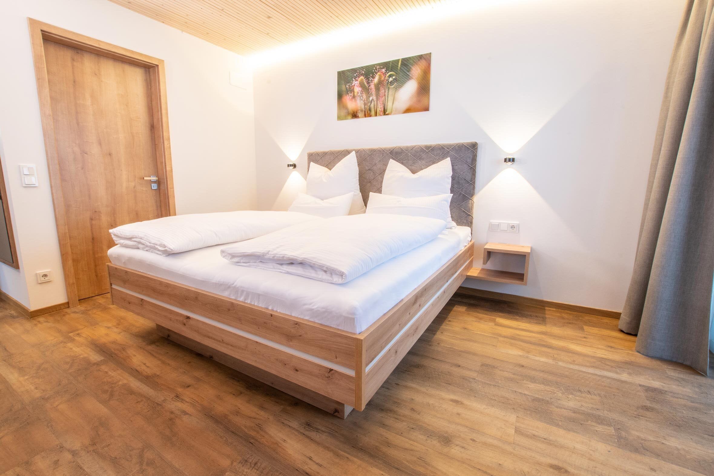 Uma cama de casal com estrutura de madeira, roupa de cama branca e cabeceira com padrão de diamante. Há uma pequena mesa de cabeceira e uma luminária fixada na parede.