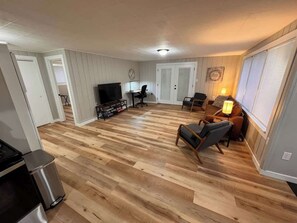 A spacious living room with a wooden floor, a television, a dining area, and a kitchen area. - Spacious Work Friendly Cottage (Bremerton)