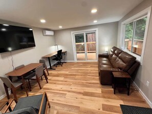 Amplia sala de estar con piso de madera, un sofá de cuero marrón, una mesa de comedor con sillas, un televisor de pantalla plana montado en la pared y una puerta corrediza de vidrio que da a una terraza exterior.