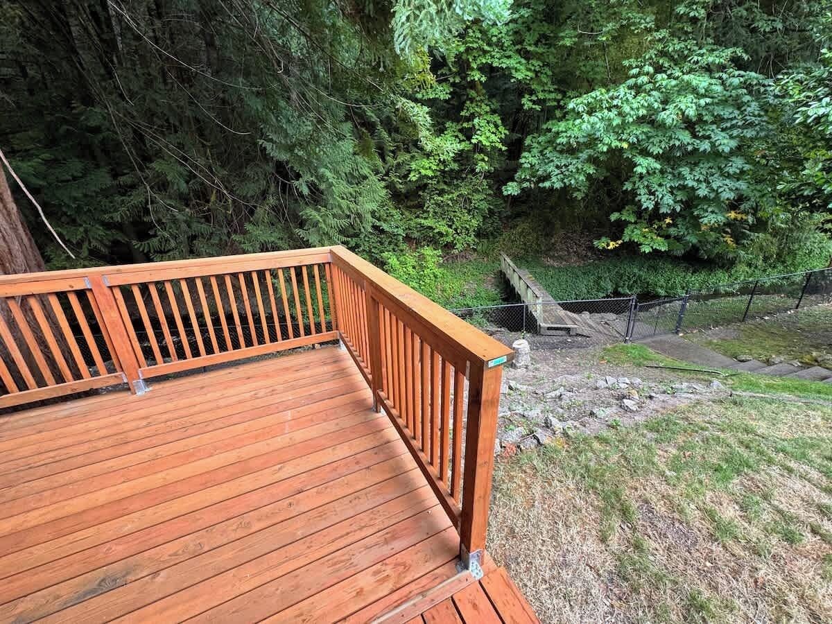 A wooden deck with a railing, surrounded by dense greenery and a stone wall.