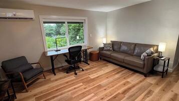 A room with a wooden floor, a desk with a chair, a sofa, and a window with a view of greenery.