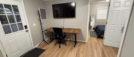 Ein Zimmer mit Holzboden, einem Schreibtisch mit Stuhl, einem an der Wand befestigten Fernseher und einer Tür zu einem weiteren Raum.