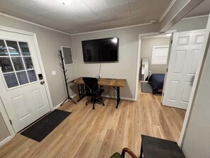 Ein Zimmer mit Holzboden, einem Schreibtisch mit Stuhl, einem an der Wand befestigten Fernseher und einer Tür zu einem weiteren Raum.