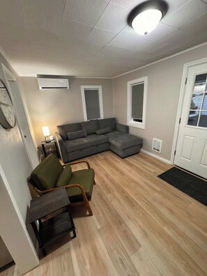 Ein Wohnzimmer mit einem Ecksofa, einem kleinen Tisch und einem Sessel. Zwei Fenster mit Jalousien, eine Tür und ein Klimagerät an der Wand sind zu sehen.