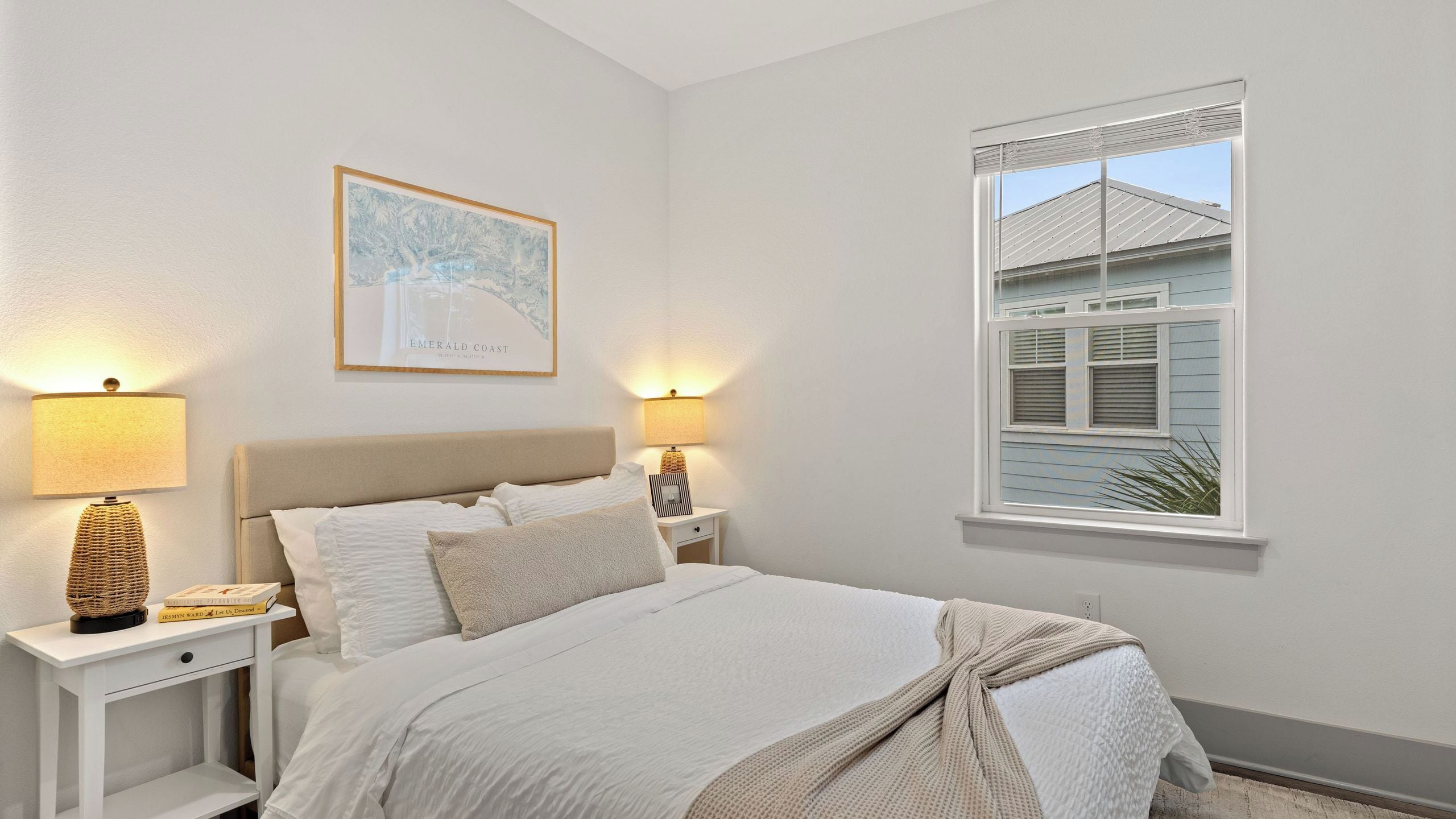 Une chambre à coucher avec un lit, deux tables de chevet, une commode, un tableau encadré et une fenêtre donnant sur une maison.