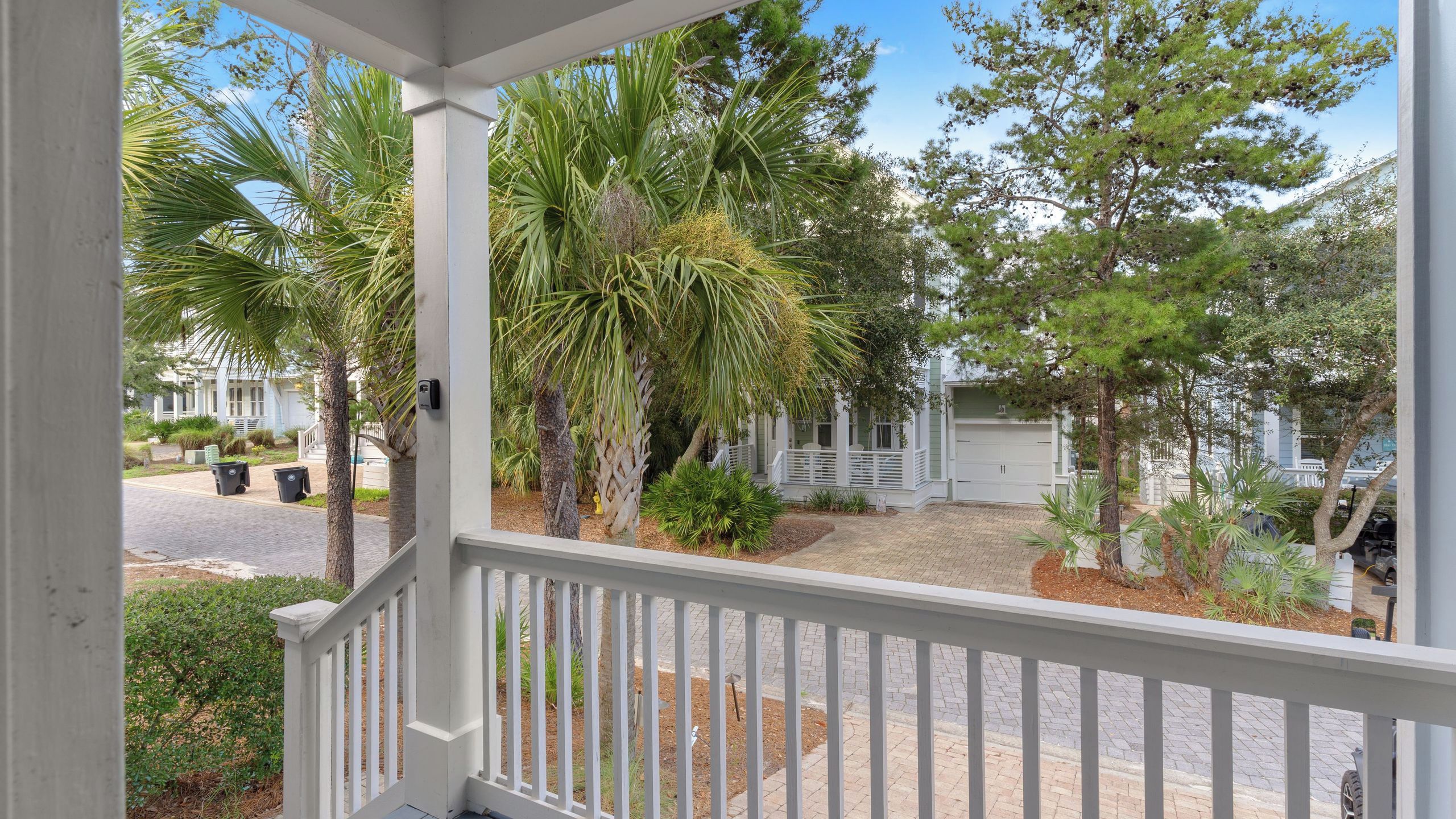 A covered outdoor area with a white railing, surrounded by various trees and shrubs, leading to a building with a white garage.